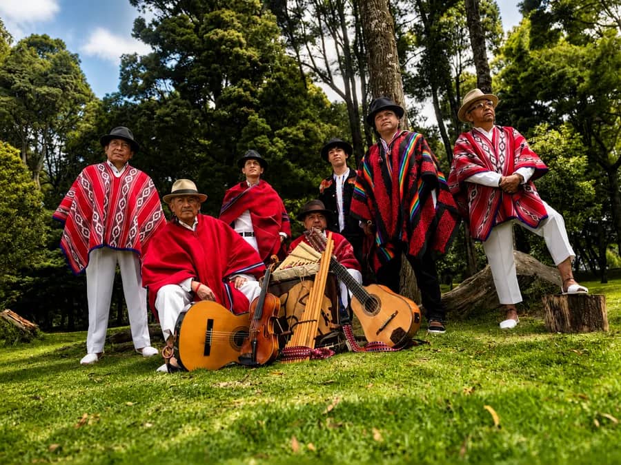 Seven members of Kapary gathered outdoors with Andean string, wind, and percussion instruments